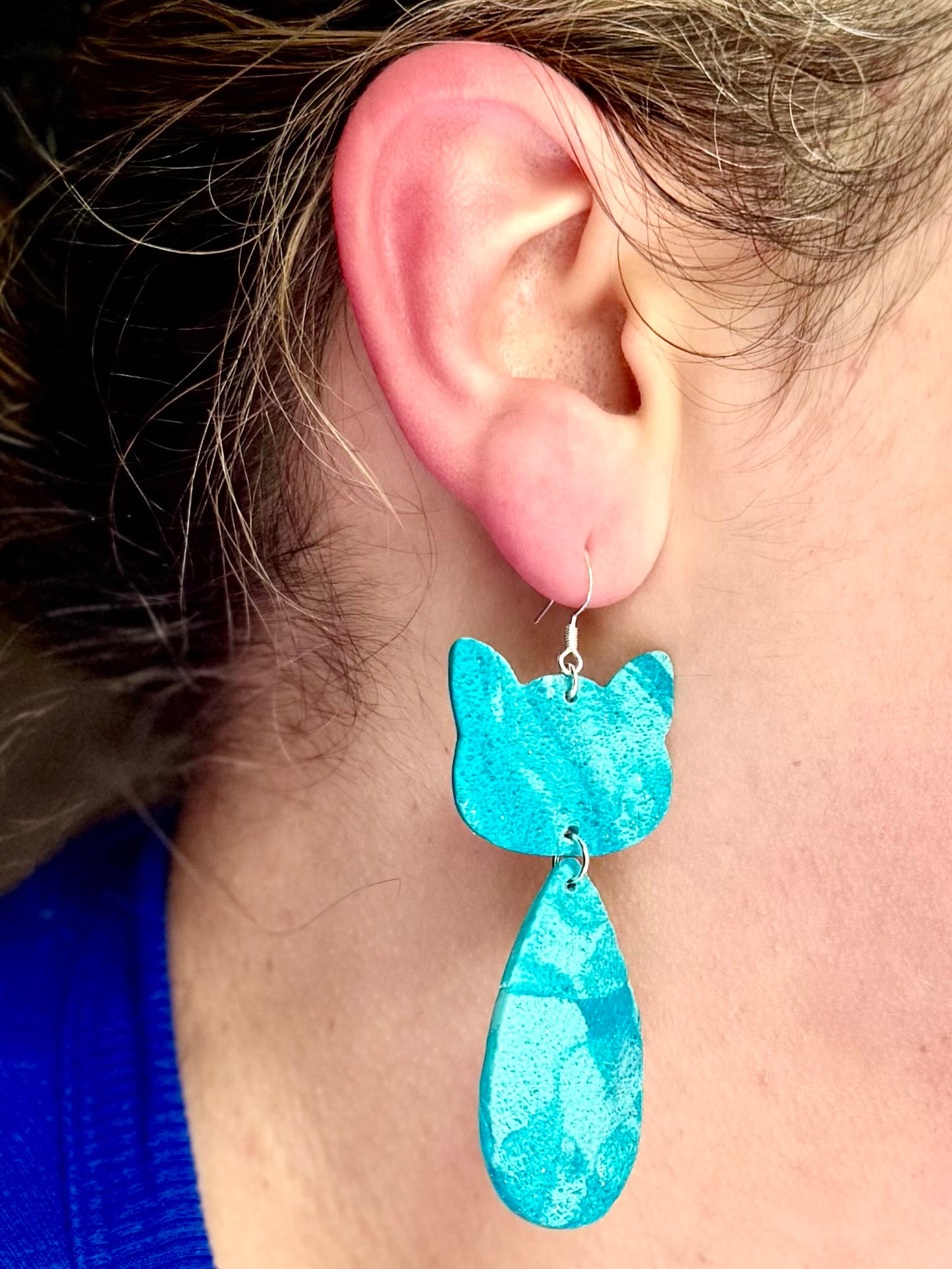 Handmade polymer clay dangle earrings in a teardrop shape with a bold marbled pattern of deep teal and light blue. Shown on a model's ear.