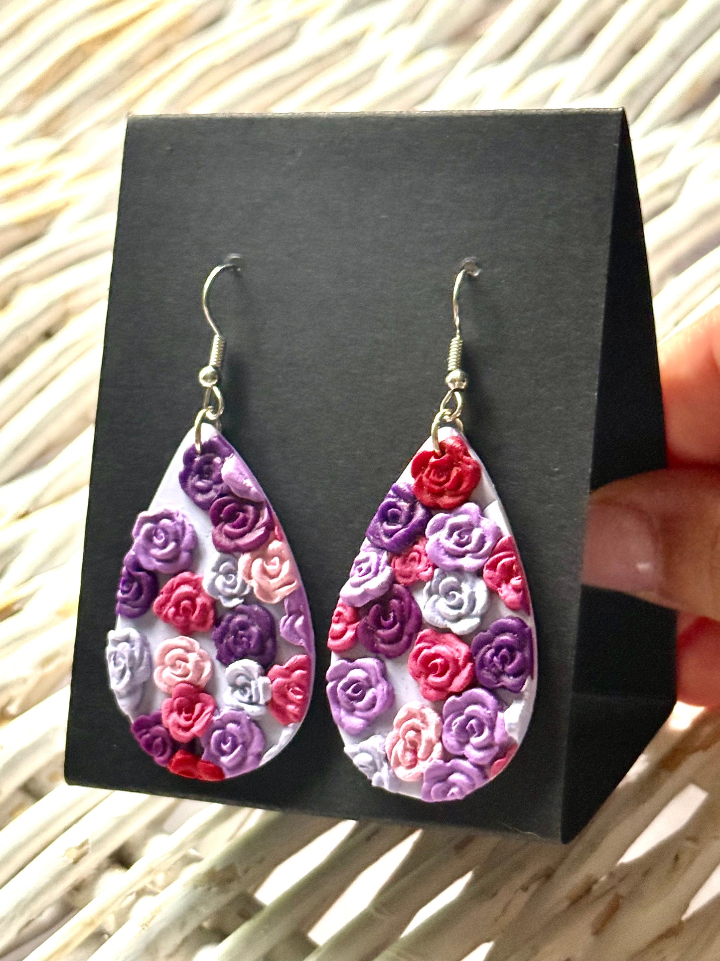 Handmade polymer clay dangle earrings in an oval shape with a delicate, painted floral pattern in pastel pinks and purples. They are displayed on a black earring card.