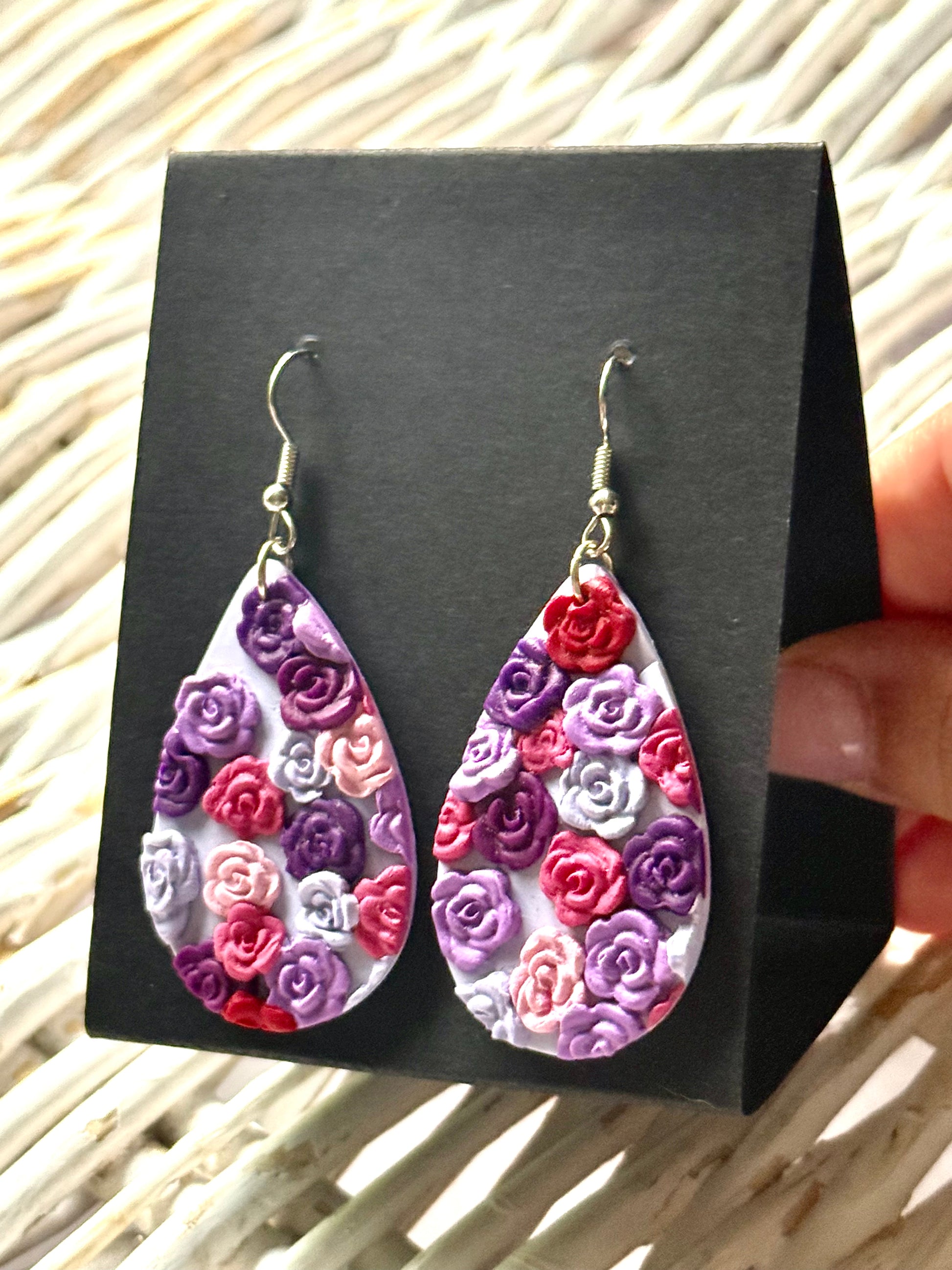 Handmade polymer clay dangle earrings in an oval shape with a delicate, painted floral pattern in pastel pinks and purples. They are displayed on a black earring card.