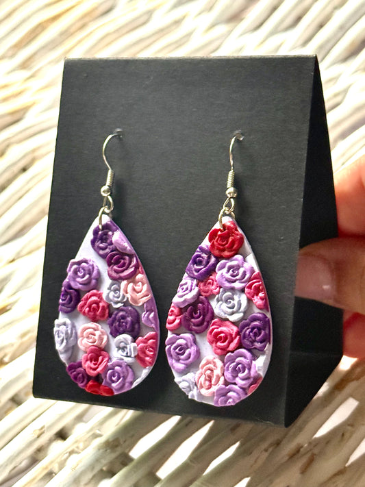Handmade polymer clay dangle earrings in an oval shape with a delicate, painted floral pattern in pastel pinks and purples. They are displayed on a black earring card.